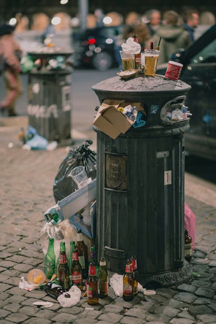 A black, cylindrical rubbish bin made of metal with a ribbed texture, standing on a cobblestone sidewalk, is shown in the image. The bin has an open slot at the top, with the lid removed or pushed aside, revealing partial contents inside. Several crumpled paper cups, some containing remnants of drinks, are placed on top of the bin, including a clear plastic cup with a straw, a paper cup with foam and a straw, and a small red disposable cup. Around the base of the bin, there is a collection of spilled and discarded rubbish, including multiple glass bottles — some green, some brown — and a clear plastic container with residual content. A black garbage bag is tied and leaning against the side of the bin, with additional litter and paper scattered on the cobblestones. In the blurred background, pedestrians are visible on a busy street, with parked cars and a grey rubbish container further back, contributing to a representation of urban rubbish accumulation and the need for regular waste collection, as addressed by services like those offered by rubbishclearanceelephantandcastle.com in managing local waste outside of municipal collections.