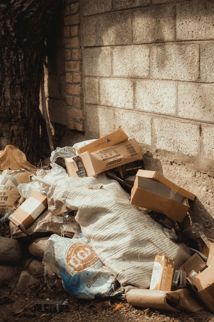 A pile of dumped waste consisting of cardboard boxes, plastic bags, and packaging materials gathered at the base of a brick wall in an outdoor area. The cardboard boxes are various sizes, some flattened and others partially intact, with visible printed labels and logos, showing signs of crumpling and used condition. The plastic bags are filled with unknown contents and appear slightly dirty or torn, with some of the packaging spilling onto uneven ground composed of gravel and small stones. In the background, a pale-colored, rough-textured brick wall rises vertically, with a dark tree trunk partially visible on the left edge, suggesting an outdoor setting near a building or fence. The scene is illuminated by natural daylight, creating soft shadows and a neutral, environmental atmosphere, which ties into the context of private rubbish removal or independent waste collection services such as those offered by rubbishclearanceelephantandcastle.com, highlighting the need for professional rubbish clearance to manage waste disposal efficiently and responsibly.