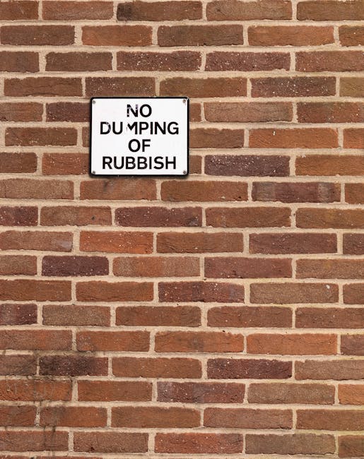 A white rectangular sign with black text reading 'NO DUMPING OF RUBBISH' is mounted on a brick wall. The wall consists of uniform, reddish-brown bricks with a rough texture, arranged in a typical running bond pattern, separated by light-colored mortar joints. The sign is positioned centrally on the upper left side of the image, and the brick surface shows slight variation in color and texture. The lighting appears natural, casting subtle shadows on the bricks, with no visible surrounding clutter or objects other than the wall and the sign. This scene reflects an outdoor urban environment where clear signage such as this might indicate a private or designated area, relevant to rubbish removal or waste management services like those provided by rubbishclearanceelephantandcastle.com, emphasizing the importance of obeying local waste disposal regulations and supporting independent collection efforts.