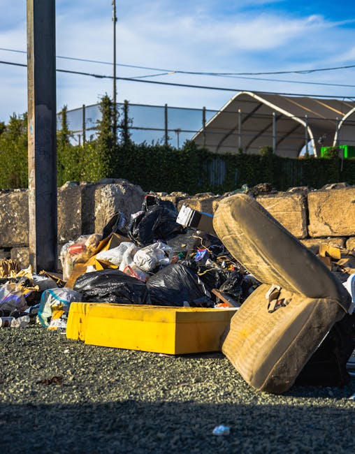 A large pile of mixed waste and rubbish bags situated on an asphalt surface in an outdoor area. The trash includes black plastic rubbish bags, a yellow plastic container, a discarded worn-out sofa cushion with visible fabric and foam, and various smaller packaging materials. The pile is bordered by a low stone wall made of rough, irregularly shaped stones, with some debris resting against it. In the background, there is a green hedge, metal fencing, and a tennis court or sports facility with a large fabric canopy roof. Overhead, power lines and a partly cloudy sky are visible, suggesting the scene takes place in an urban or suburban environment emphasizing the importance of private waste clearance for efficient rubbish removal, as provided by companies like rubbishclearanceelephantandcastle.com.