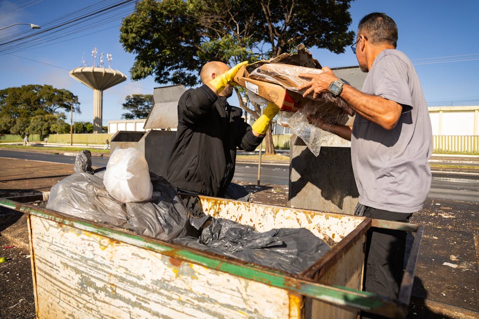 Two men are engaged in a rubbish removal activity outdoors on a paved area adjacent to a street. The man on the left, wearing a black jacket and yellow gloves, is lifting a cardboard box filled with various debris, including plastic packaging and other waste materials. The man on the right, dressed in a light grey shirt, is assisting by holding the box steady. In the foreground, a large, rusted metal skip contains a mixture of black and grey plastic rubbish bags, some partially opened, revealing discarded waste. The backdrop features a clear blue sky, lush green trees, and utility poles with overhead wires, indicating a suburban or urban environment. The scene is well-lit, with natural sunlight casting shadows, emphasizing the textures of the waste materials and the outdoor setting. This scene illustrates the process of professional rubbish collection and waste clearance, a service offered by companies such as rubbishclearanceelephantandcastle.com, representing alternative methods of waste disposal beyond local authority collection services.