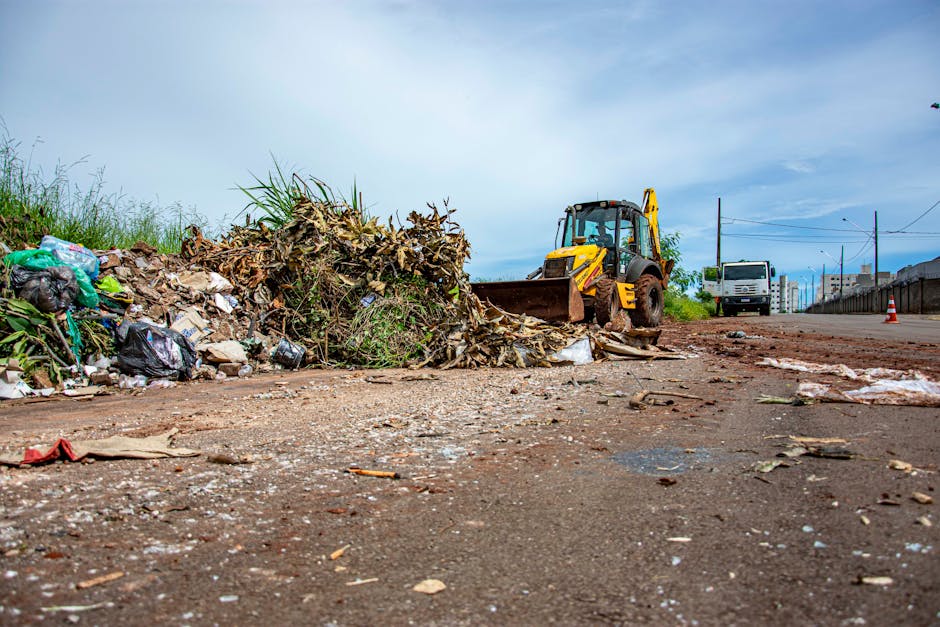 A scene depicting roadside waste clearance with a yellow backhoe loader positioned behind a large pile of mixed rubbish. The pile contains various waste materials including plastic bags, paper, and plant debris, which are loosely heaped on the ground and partially on the edge of the asphalt road. The surface of the road appears worn and slightly uneven, with small scattered debris and grime visible in the foreground. In the background, there is a white van parked further along the road, along with a construction cone and utility poles with power lines overhead, indicating an area of ongoing work or disturbance. The environment is outdoors under a partly cloudy sky, and the setting suggests a local effort for the removal and management of bulky waste, reflecting a form of independent or private disposal rather than council collection, with firm visual cues linking it to waste management services such as those offered by rubbishclearanceelephantandcastle.com.