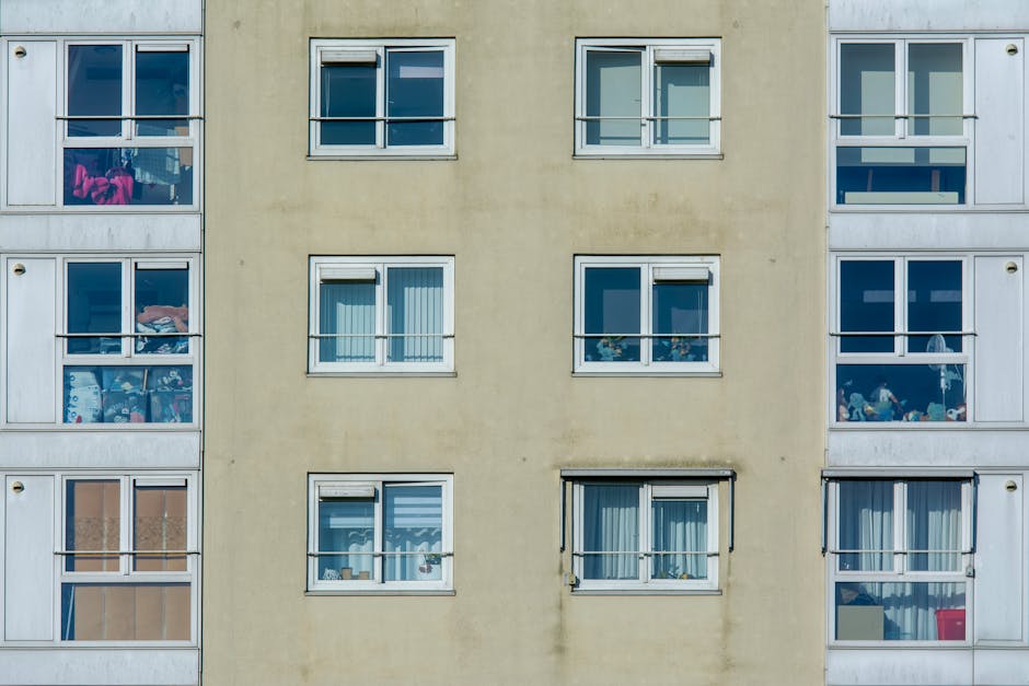 A close-up view of the exterior of a multi-storey residential building showing a yellow central section flanked by two white-framed sections. The yellow wall features six evenly spaced rectangular windows with white trims, some with partially drawn blinds or curtains. The left white section contains several balconies filled with various items such as clothing, laundry, and household objects, indicating different levels of outdoor space utilization. The right white section also has balconies, which are decorated with toys, plants, and personal belongings, with some glass panels reflecting light. The overall scene depicts an urban living environment with visible signs of daily household activity and personalized balcony spaces, characteristic of apartment building facades. This background can be associated with urban waste management scenarios, including private rubbish collection or on-site clearance for lease or renovation purposes, which are common in such residential settings, as handled by rubbish clearance services like those provided by rubbishclearanceelephantandcastle.com.