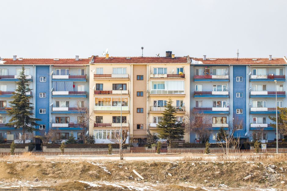 A close-up view of the exterior of a multi-storey residential building showing a yellow central section flanked by two white-framed sections. The yellow wall features six evenly spaced rectangular windows with white trims, some with partially drawn blinds or curtains. The left white section contains several balconies filled with various items such as clothing, laundry, and household objects, indicating different levels of outdoor space utilization. The right white section also has balconies, which are decorated with toys, plants, and personal belongings, with some glass panels reflecting light. The overall scene depicts an urban living environment with visible signs of daily household activity and personalized balcony spaces, characteristic of apartment building facades. This background can be associated with urban waste management scenarios, including private rubbish collection or on-site clearance for lease or renovation purposes, which are common in such residential settings, as handled by rubbish clearance services like those provided by rubbishclearanceelephantandcastle.com.